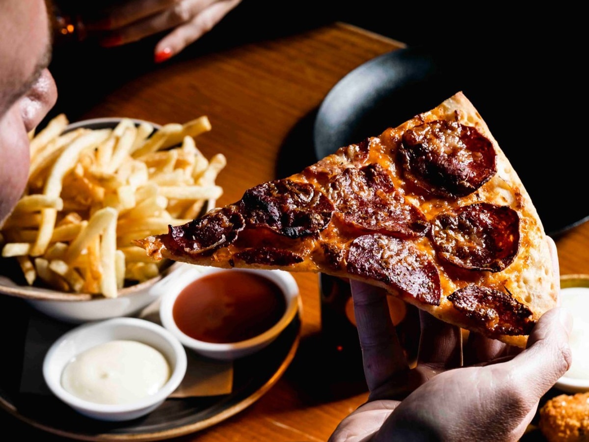 man holding slice of pizza with bowl of fries and dips on the table